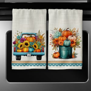 May include: Two kitchen towels with a fall theme. One towel features a blue pickup truck filled with pumpkins and fall flowers. The other towel features a teal bucket filled with pumpkins and fall flowers.