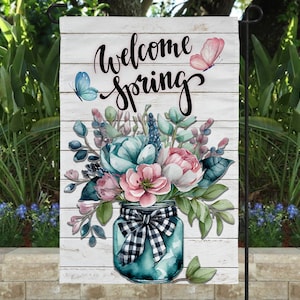 May include: Welcome Spring garden flag with a white wood background. A mason jar filled with a bouquet of pink, blue, and green flowers with butterflies on the top. The jar has a black and white checkered ribbon.