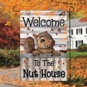 May include: A welcome garden flag with a squirrel peeking out from behind a wooden fence. The flag has a white background with brown wood planks and autumn leaves. The text on the flag reads "Welcome to the Nut House".