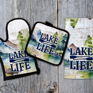 Puede incluir: Un conjunto de tres accesorios de cocina con un diseño de acuarela de una escena de lago con árboles y montañas. El conjunto incluye un posavasos, un guante para horno y una toalla de cocina. Cada artículo presenta el texto "Lake Life" con dos remos cruzados.