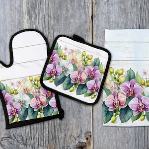 May include: A set of three kitchen accessories featuring a floral design with white and purple orchids on a whitewashed wood background. The set includes a pot holder, oven mitt, and a tea towel.