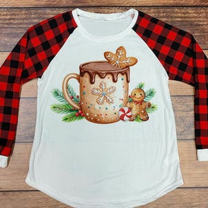 Può includere: Maglietta bianca a maniche lunghe con maniche a quadri rossi e neri. La parte anteriore presenta una grafica di una tazza di cioccolata calda con un biscotto di pan di zenzero, un omino di pan di zenzero e un bastoncino di zucchero.