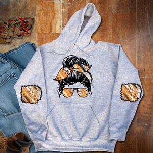 May include: A gray hoodie with a black and white illustration of a woman with a messy bun and sunglasses. The bun is decorated with a patterned orange and brown ribbon. The hoodie has matching patterned patches on the elbows.