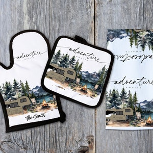 Puede incluir: Un juego de tres textiles de cocina con una ilustración de acuarela de una autocaravana estacionada en un entorno boscoso con una fogata y montañas en el fondo. El texto "Adventure Awaits" está escrito en la toalla y los agarraderas. El agarradera tiene el texto "The Senas" en la parte inferior.