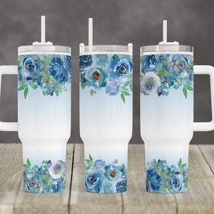 May include: White insulated tumbler with a handle, featuring a watercolor floral design in shades of blue and green. The floral pattern is arranged at the top and bottom of the tumbler, with a light blue gradient in the center. Includes a white lid and straw.