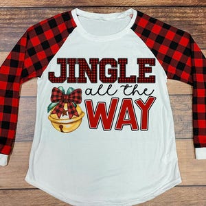 Könnte beinhalten: Weißes Langarmshirt mit rot-schwarz karierten Ärmeln. Auf der Vorderseite steht der Text "JINGLE all the WAY" und eine Weihnachtsglocken-Grafik. Ein festliches Urlaubsdesign.