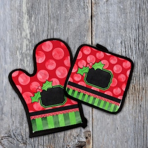 Op de afbeelding: Een rode ovenwant en pannenlap set met een kerstthema. De set heeft een rood stippenpatroon met een zwart schoolbordontwerp, hulstaccenten en groen en rood gestreepte rand.