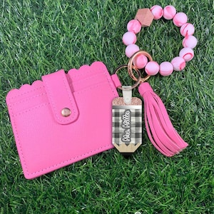 May include: A pink leather card holder with a scalloped edge and a snap closure. A black and white checkered pencil keychain with the text "Mrs. Miller" is attached to a pink beaded bracelet with a gold charm.