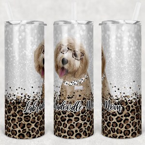 May include: Three stainless steel tumblers with a glitter background and a leopard print design. The center tumbler features a doodle dog wearing glasses and a collar with a bone charm. The text "Doodle Mom" is printed on the tumbler. The left tumbler has the text "Labradoodle" and the right tumbler has the text "Mom".