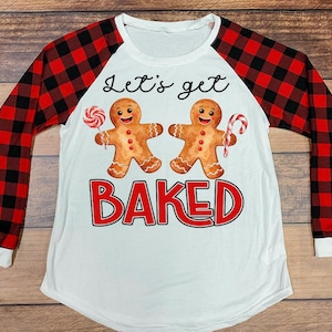 Könnte beinhalten: Weißes Langarmshirt mit rot-schwarz karierten Ärmeln. Das Shirt zeigt zwei Lebkuchenmänner mit Süßigkeiten und den Text "Let's get BAKED" in Schwarz und Rot.