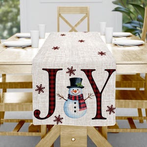 May include: A beige table runner with the word "JOY" in large, red plaid letters. A snowman with a black top hat and red plaid scarf is in the center. Red snowflakes are scattered across the runner. The runner is on a wooden table.