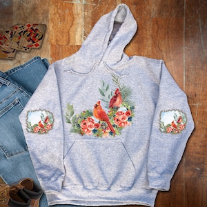 May include: A gray hooded sweatshirt with a floral design featuring three red cardinals. The design is centered on the front of the sweatshirt and includes a variety of flowers and greenery. The sweatshirt has a pocket on the front and the sleeves have a smaller floral design with a cardinal on each sleeve.