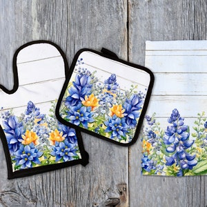 Peut inclure: Un ensemble de trois textiles de cuisine avec un motif floral de bleuets sur un fond de bois blanchi à la chaux. L'ensemble comprend un dessous de plat, un gant de cuisine et une serviette à thé.