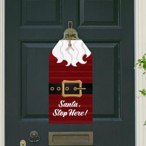Op de afbeelding: Een kerstdeurhanger in de vorm van de outfit van de kerstman. Het rode houten bord heeft een witte baard, een zwarte riem met een gouden gesp en de tekst "Santa, Stop Here!" in het wit. Er is een jute touw bevestigd om op te hangen.