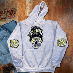 May include: Gray hoodie with a black and white skull design with yellow and black stripes on the sleeves. The skull has a messy bun with a yellow and black striped bow.