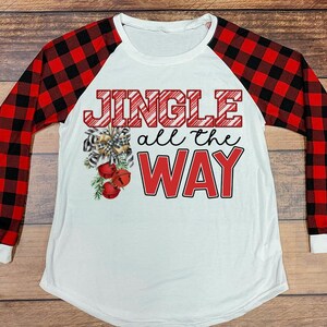Könnte beinhalten: Weißes Langarmshirt mit rot-schwarz karierten Ärmeln. Das Shirt zeigt den Text "JINGLE all the WAY" in Rot, zusammen mit einer festlichen Grafik von Glocken und einer Schleife. Ein Oberteil zum Thema Feiertage.