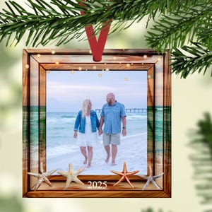 May include: Square wooden Christmas ornament with a photo of a couple walking on a beach. The ornament has a wooden frame with a red ribbon for hanging. The year 2025 is printed at the bottom, and starfish are placed at the bottom of the frame.