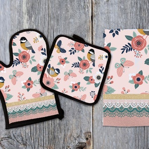 May include: A set of three pink kitchen accessories with a floral and bird pattern. The set includes a pot holder, oven mitt, and a tea towel. Each item has a lace trim and a gold accent.