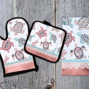 May include: A set of three kitchen accessories featuring a white background with a pattern of colorful, stylized sea turtles. The set includes a pot holder, oven mitt, and a tea towel. All three items have a pink and white lace trim.
