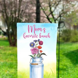 Puede incluir: Una bandera de jardín azul y blanca con un fondo de acuarela. La bandera presenta una lata de leche blanca con un asa de madera llena de un ramo de flores rosas, rojas, moradas y azules. El texto "Mom's favorite bunch" está escrito en letras cursivas rosas.