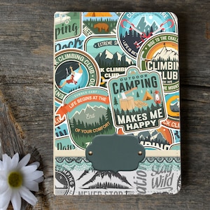May include: A colorful notebook with a variety of camping and climbing themed stickers. The stickers feature images of mountains, trees, tents, axes, and other outdoor adventure elements. The notebook has a green label with a blank space for writing.