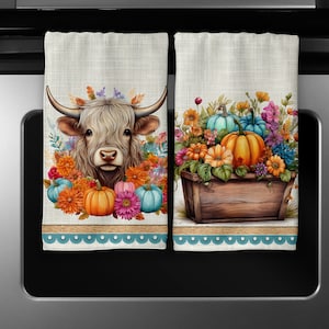 May include: Two kitchen towels with an autumnal theme. One towel features a white highland cow with a floral wreath around its head. The other towel features a wooden box overflowing with pumpkins and flowers.