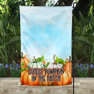 Op de afbeelding: Een tuinvaandel met een blauwe luchtachtergrond en de tekst "Cutest Pumpkin in the Patch". De vlag toont aquarel pompoenen in tinten oranje en wit, met groene bladeren en ranken. Een decoratief item voor de herfst.