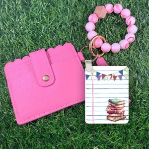 May include: Pink leather card holder with a scalloped edge and a snap closure. A white card with a stack of books and a banner of colourful flags is attached to a pink and white beaded bracelet.