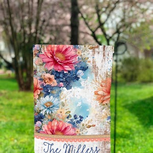 Puede incluir: Una bandera de jardín con un diseño floral con flores rosas, azules y amarillas sobre un fondo blanco con textura de madera desgastada. La bandera tiene un borde marrón con un borde festoneado blanco y el texto "The Millers" en negro.