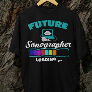 Puede incluir: Camiseta negra con el texto "FUTURE Sonographer LOADING..." en turquesa y blanco. El diseño incluye un portátil con una imagen de ultrasonido y una barra de carga con segmentos coloridos. La camiseta está colgada en una percha de madera.