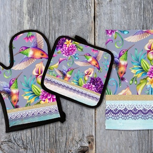 May include: A set of three kitchen accessories featuring a hummingbird and floral pattern. The set includes a potholder, an oven mitt, and a tea towel. All three items have a gray background with colorful hummingbirds and flowers. The edges of the items are trimmed with white lace.