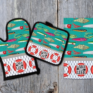 Pode incluir: Um conjunto de três acessórios de cozinha com um fundo turquesa com um padrão de iscas de pesca e peixes. O conjunto inclui um descanso de panela, uma luva de forno e um pano de prato. Cada item tem um salva-vidas vermelho e branco com o texto "Lake Life" em preto.