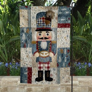 May include: A decorative garden flag featuring a quilted design of a nutcracker soldier. The flag has a patchwork background with blue, red, and cream-colored squares. The nutcracker is dressed in a blue and red uniform with a black mustache and beard.
