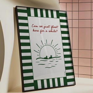 May include: A framed print with a green and white striped border. The print features a white background with a green silhouette of a sunset and two people in a boat. The text "Can we just float here for a while?" is written in red.