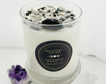 May include: A white soy wax candle in a clear glass jar with a black label that reads "100% Natural Soy Wax" and "Hand Poured in Brooklyn, NY". The candle is topped with black and silver glitter and a black stone.