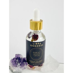 May include: A glass bottle with a gold lid and a dropper top. The bottle is filled with a clear liquid and contains dried flowers. The label on the bottle reads "Libra Goddess Zodiac Moon Child Essential Oil Blend Lavender, Frankincense, Cedar, Orange & Rose Net Wt 3 oz."