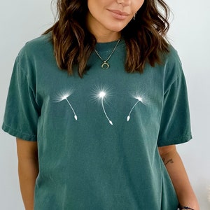 May include: A dark teal blue t-shirt with a white graphic of three dandelions blowing in the wind. The shirt has a relaxed fit and a crew neck.