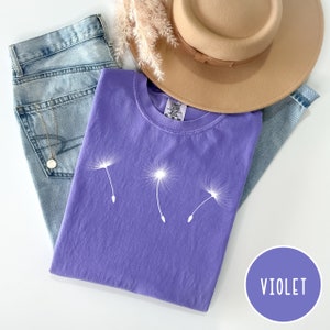 May include: A purple t-shirt with a white dandelion design. The shirt is laying on a pair of blue jeans and a brown hat. The word "VIOLET" is written in a circle on a purple background.