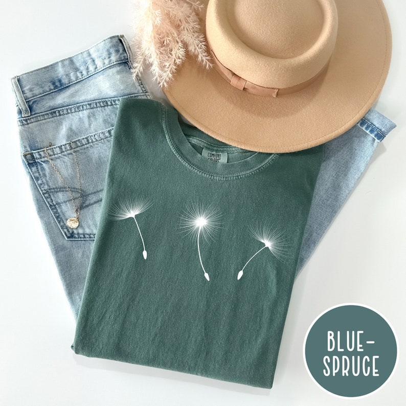 May include: A green t-shirt with a white dandelion graphic. The shirt is folded and laid on a white surface with a pair of blue jeans and a brown hat.