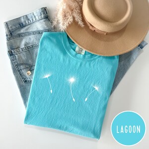 May include: A turquoise blue t-shirt with a white dandelion graphic. The t-shirt is folded and laid on a light blue denim background with a brown hat and a light brown dried flower arrangement.