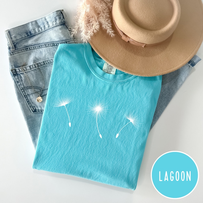 May include: A turquoise blue t-shirt with a white dandelion graphic. The t-shirt is folded and laid on a light blue denim background with a brown hat and a light brown dried flower arrangement.