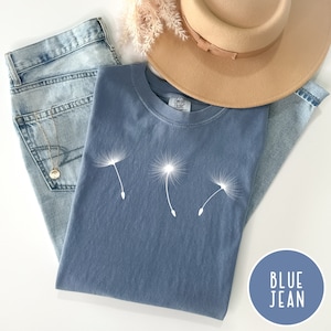 May include: A blue jean t-shirt with a white graphic of three dandelions blowing in the wind. The shirt is laid flat on a white surface with a pair of blue jeans and a brown hat.