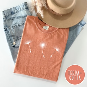 May include: A terracotta colored t-shirt with a white dandelion graphic. The shirt is laying on a pair of blue jeans and a brown hat.