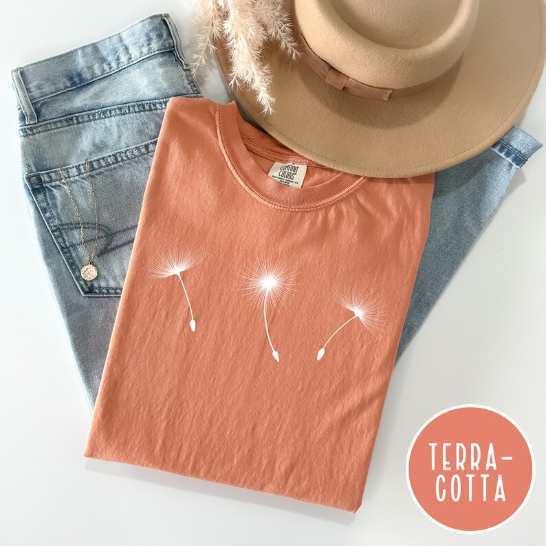 May include: A terracotta colored t-shirt with a white dandelion graphic. The shirt is laying on a pair of blue jeans and a brown hat.