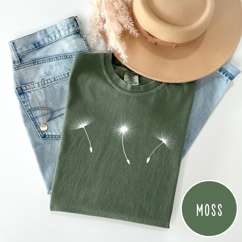 May include: A green t-shirt with a white dandelion graphic. The shirt is folded and laid on a white surface with a pair of blue jeans and a brown hat. The color of the shirt is labeled "Moss".