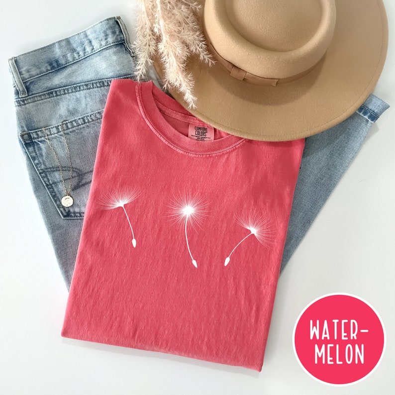 May include: A red t-shirt with a white dandelion graphic. The shirt is folded and laid on a white surface with a pair of blue jeans and a brown hat. The text "WATER-MELON" is written in white on a red circle.