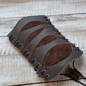 May include: A brown leather arm cuff with suede accents and a lace-up closure. The cuff is made of two different types of leather, one smooth and one suede.