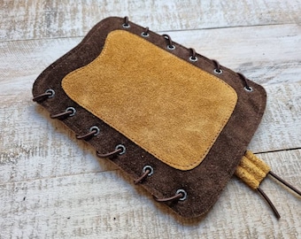 May include: A brown leather pouch with a tan suede patch and brown laces. The pouch has a flap that closes with a snap.