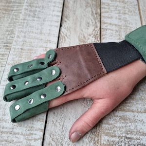 May include: A green and brown leather archery finger tab and wrist guard. The finger tab has three finger loops with silver rivets. The wrist guard is green and attached to a brown leather piece with an elastic band.