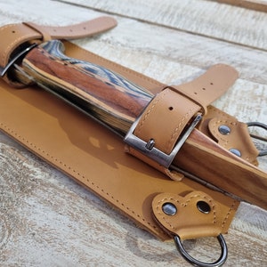 May include: A tan leather bow quiver with two straps and buckles. The quiver is designed to hold a bow and arrows. The quiver has two metal rings on the bottom.
